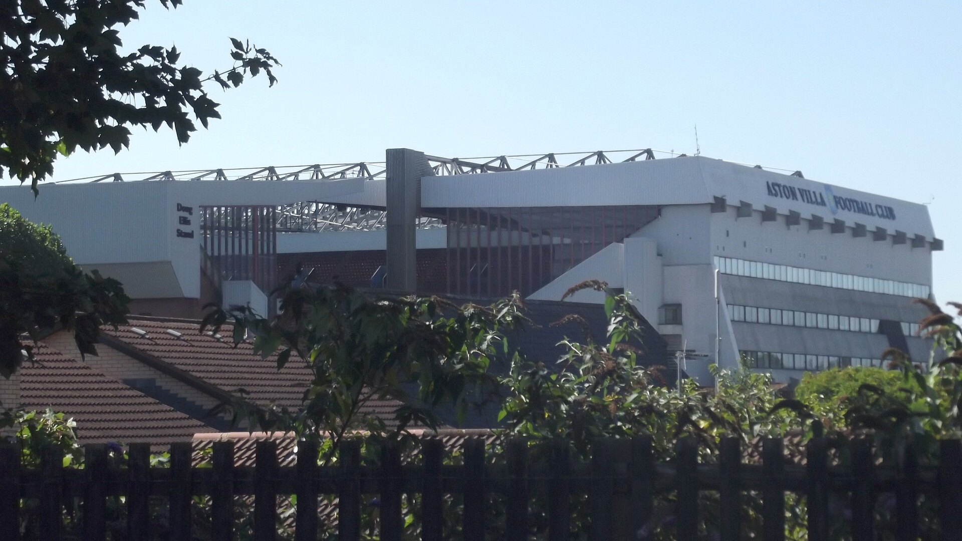 Трибуните „Дъг Елис“ и „Северна“ на стадион „Вила Парк“ с надпис Aston Villa Football Club, гледани над дървета и покриви