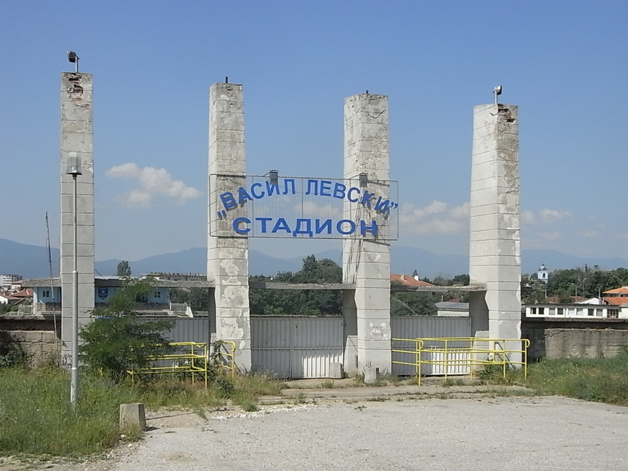 Входът на стадион „Васил Левски“ в Карлово с четири бетонни колони и синя табела, планината на фона